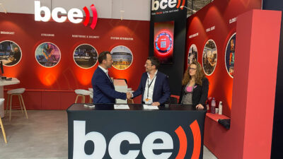 Photo prise lors de l'IBC 2022, sur le stand de BCE, capturant la signature du contrat entre BCE et LN24. À gauche, Steve De Jongh, CTO de LN24 et IPM Group, signe le contrat aux côtés de Christophe Goossens, CEO de BCE. Stéphanie Schaeffer, commerciale chez BCE, observe l'assemblée à proximité.